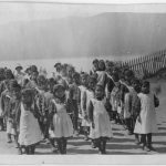 Four lines of children in school uniform marching along a path on school grounds. Ocean and mountains in the background.