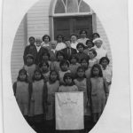 Children and adults standing posed for a photo on some steps with the auxiliary banner at centre. Image is cropped into an oval and affixed to a piece of paper.