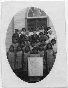 Children and adults standing posed for a photo on some steps with the auxiliary banner at centre. Image is cropped into an oval and affixed to a piece of paper.