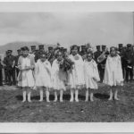 Group of six youth standing in white graduation dresses holding bouquets, larger group in uniform standing in lines behind them.