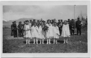 Group of six youth standing in white graduation dresses holding bouquets, larger group in uniform standing in lines behind them.