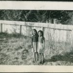 Two children with arms around another standing in yard, wood fence in the background