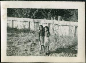 Two children with arms around another standing in yard, wood fence in the background