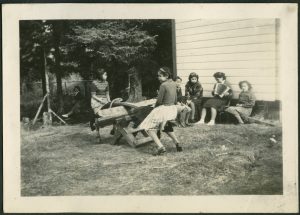 Two children are on a see-saw, one child is swinging on a swing, and there are other children sitting by a child playing the accordion