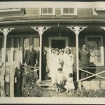Adults in formal wedding attire standing on a veranda with two small children holding flowers