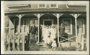 Adults in formal wedding attire standing on a veranda with two small children holding flowers