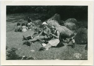 Staff and children sitting on the beach in two separate groups, crafting, staff sitting with backs against large boulders, two are without shoes, children are sitting in a semi-circle