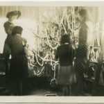 Four children decorating a Christmas tree, one is on a ladder and another on a chair decorating the top of the tree
