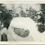 Children and staff in winter gear behind wooden structure making snowballs