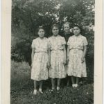 Three youth in identical dresses standing with a staff member in front of trees