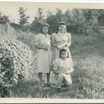 Three youth holding rolled diplomas standing next to tall hedge, one youth is kneeling