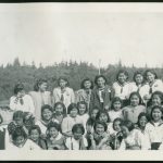 Group of youth and children on the beach, some in uniform, Port Simpson Residential School building in the distance