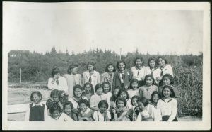Group of youth and children on the beach, some in uniform, Port Simpson Residential School building in the distance