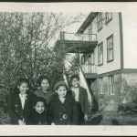 Group of children standing next to a tree with front entrance and stairs to Port Simpson Residential School in the background