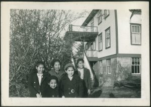 Group of children standing next to a tree with front entrance and stairs to Port Simpson Residential School in the background