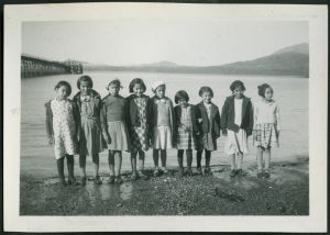 Children standing in a line on the shoreline, pier in the background
