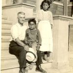 Visiting reverend posing with two children. The smaller child is seated on the reverend's lap and the other child is standing next to the reverend. They are all on the stairs of Norway House Residential School Building.