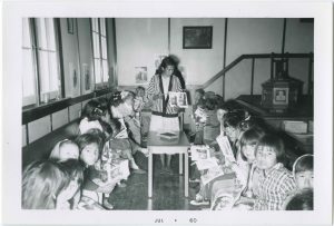 Instructor standing between two lines of younger children sitting with books, many of whom are looking at camera.